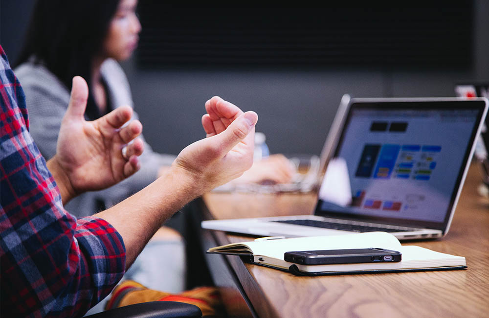 Hands gesturing in front of a laptop in the foreground and a person working on a laptop in the background.