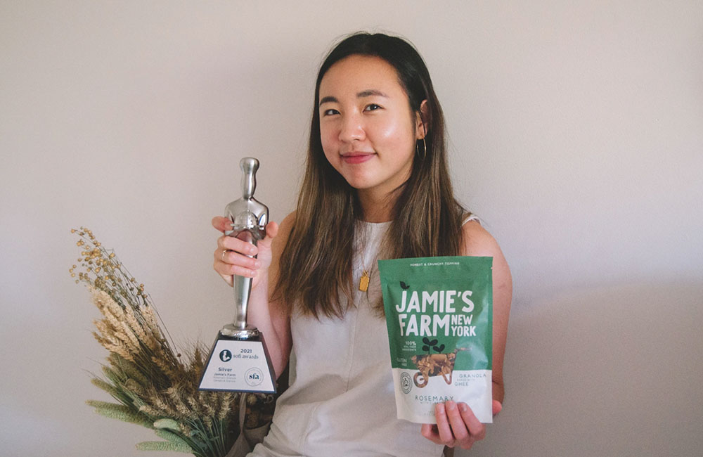 Jaime Kim, founder of Jaime's Farm, holds a trophy and a package of her granola product against a white backdrop. Behind her, strands of wheat are on display.