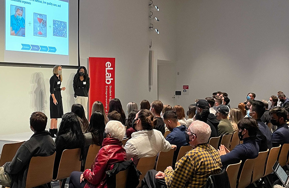 Two student pitch to an audience. A project screen displaying slides is on the wall above them and a red banner with the text "eLab" is on the stage next to them.