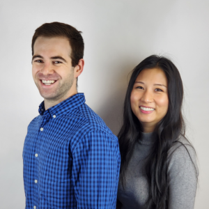 White man and asian woman stand together in front of white wall 