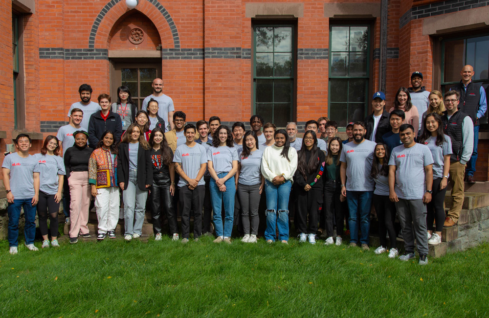 A group of college students and several professors stand outside in a grassy area in front of a building for a group photo.